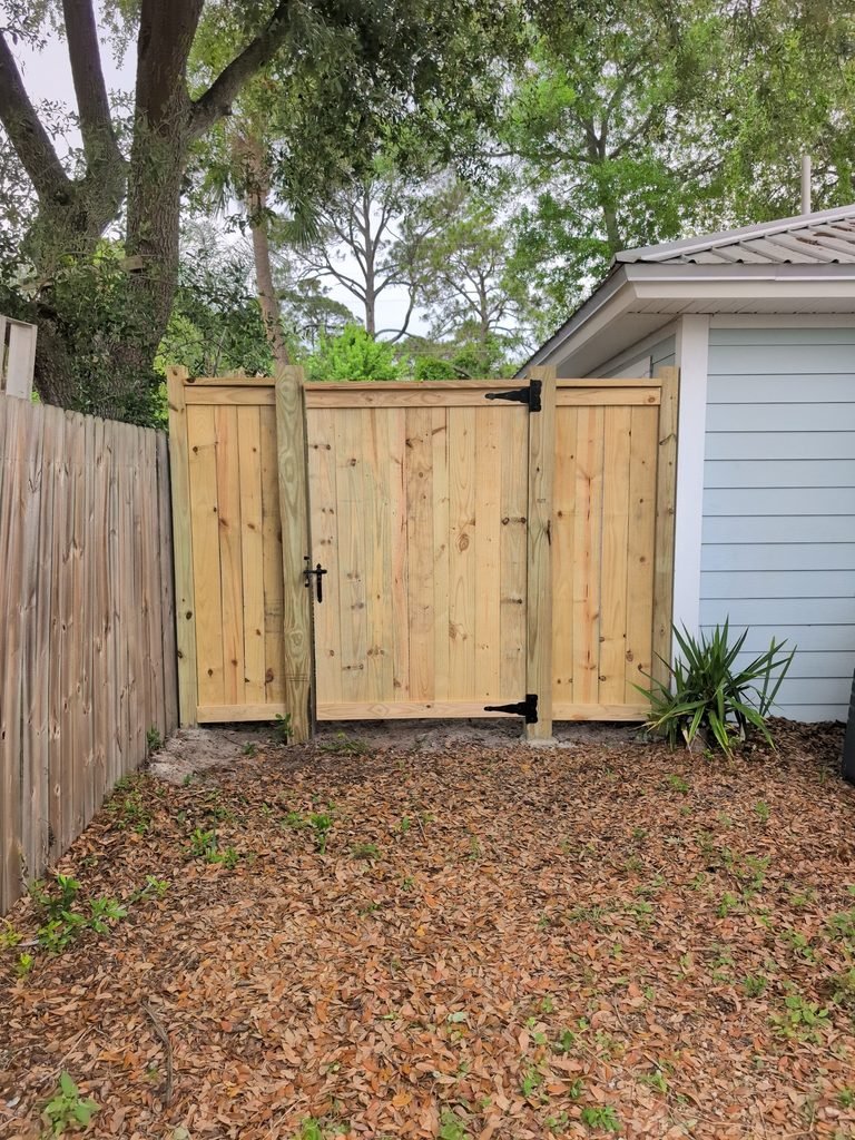 Wood privacy fence and double gate with horizontal top trim, flat-cut posts, and black hinges beside a house.